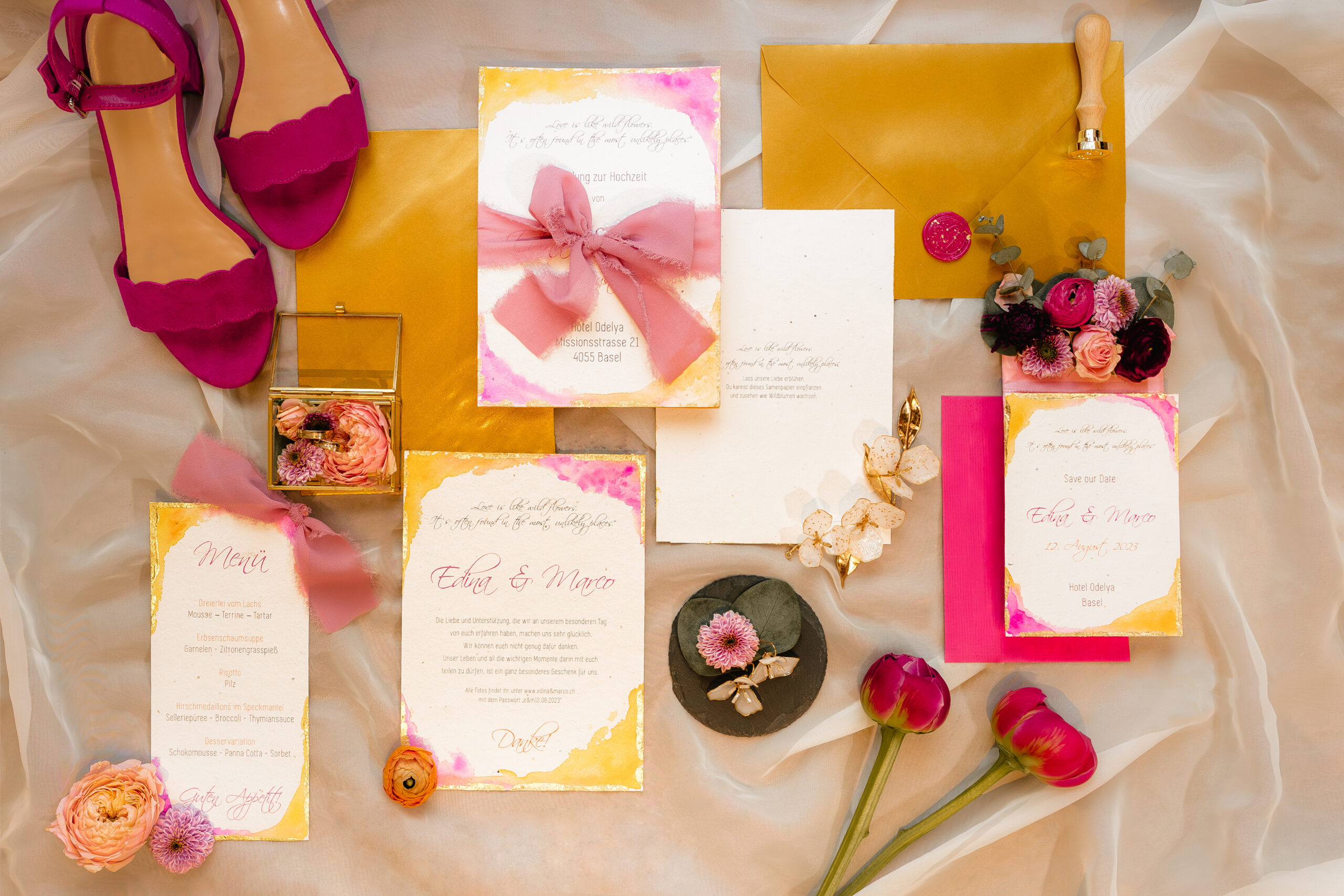 Luxuriöses Flatlay von hochwertiger Hochzeitspapeterie auf Samenpapier mit Aquarellakzenten, Blattgold & Chiffonband in Magenta und Gold